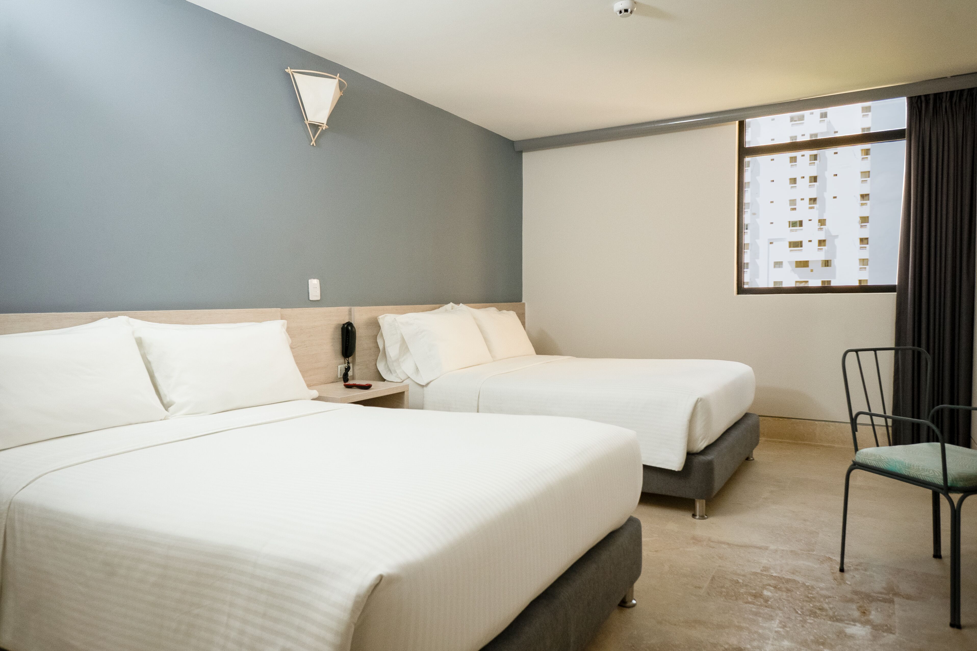 Habitación de hotel con dos camas, una silla, una mesita de noche, una lámpara de pared y un ventanal con cortinas.