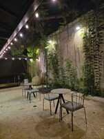 Un área de estar al aire libre con mesas y sillas, rodeada de abundante vegetación e iluminación decorativa.