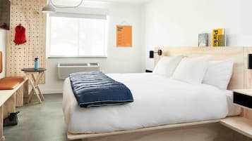 Une chambre d’hôtel moderne avec un lit en bois, un bureau avec une chaise, une petite table, un sac à dos rouge et une fenêtre avec des stores.