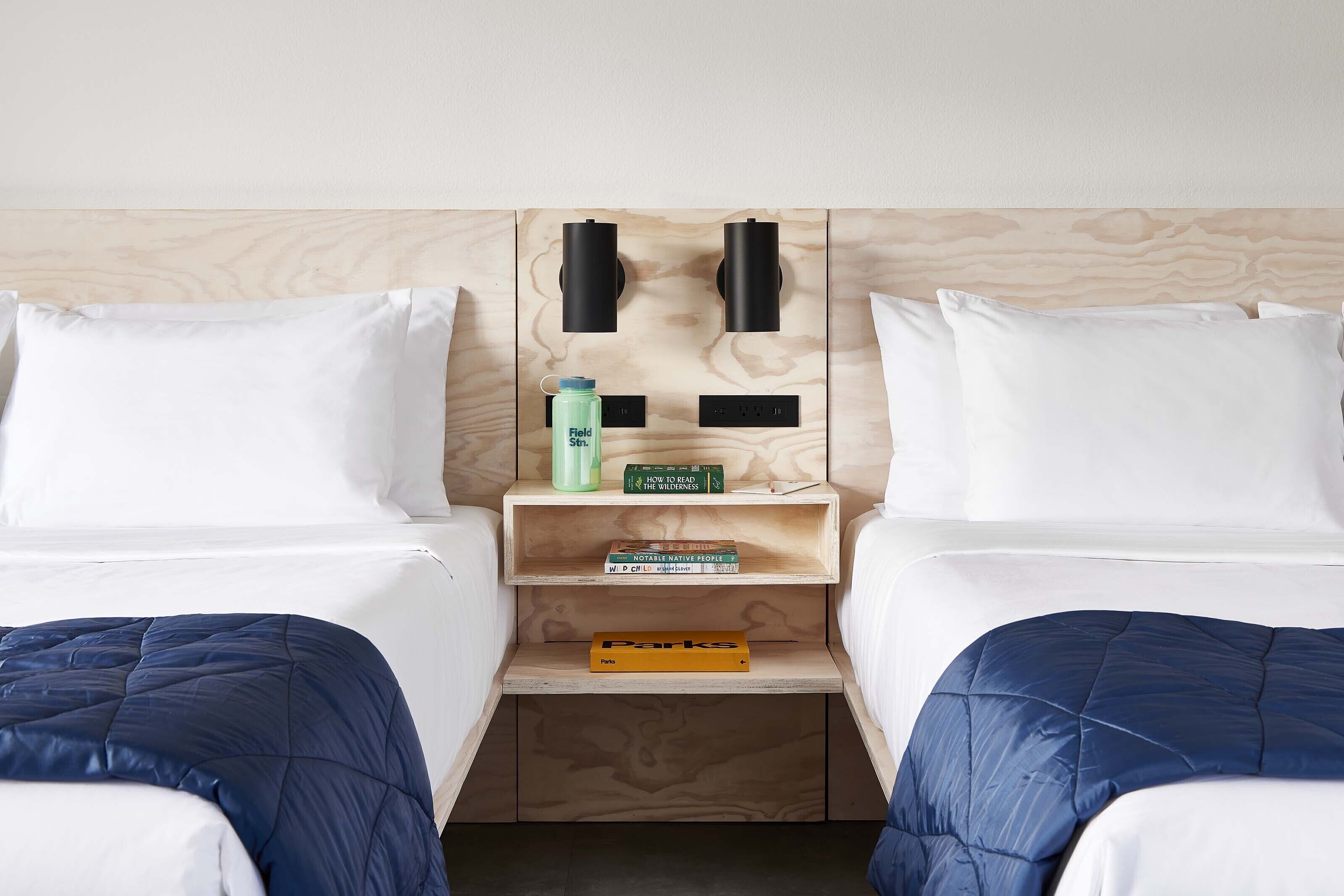A hotel room with two beds, a wooden headboard, a shelf with books, and a water bottle.
