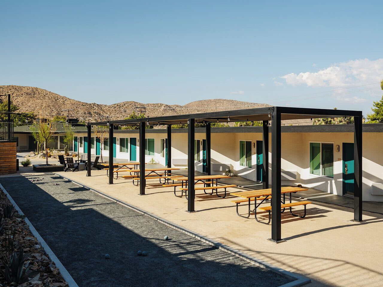 Un espacio de estar al aire libre cubierto con mesas de picnic y bancos, un edificio con ventanas y puertas, y un paisaje montañoso al fondo.