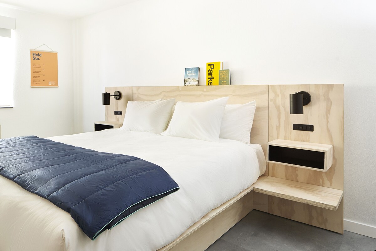 Una cama cuidadosamente hecha con colcha azul, almohadas blancas y cabecera de madera. Hay dos mesitas de noche con lámparas negras y una pequeña estantería sobre la cama con libros.