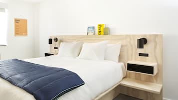 A neatly made bed with a blue quilt, white pillows, and a wooden headboard. There are two bedside tables with black lamps and a small shelf above the bed holding books.