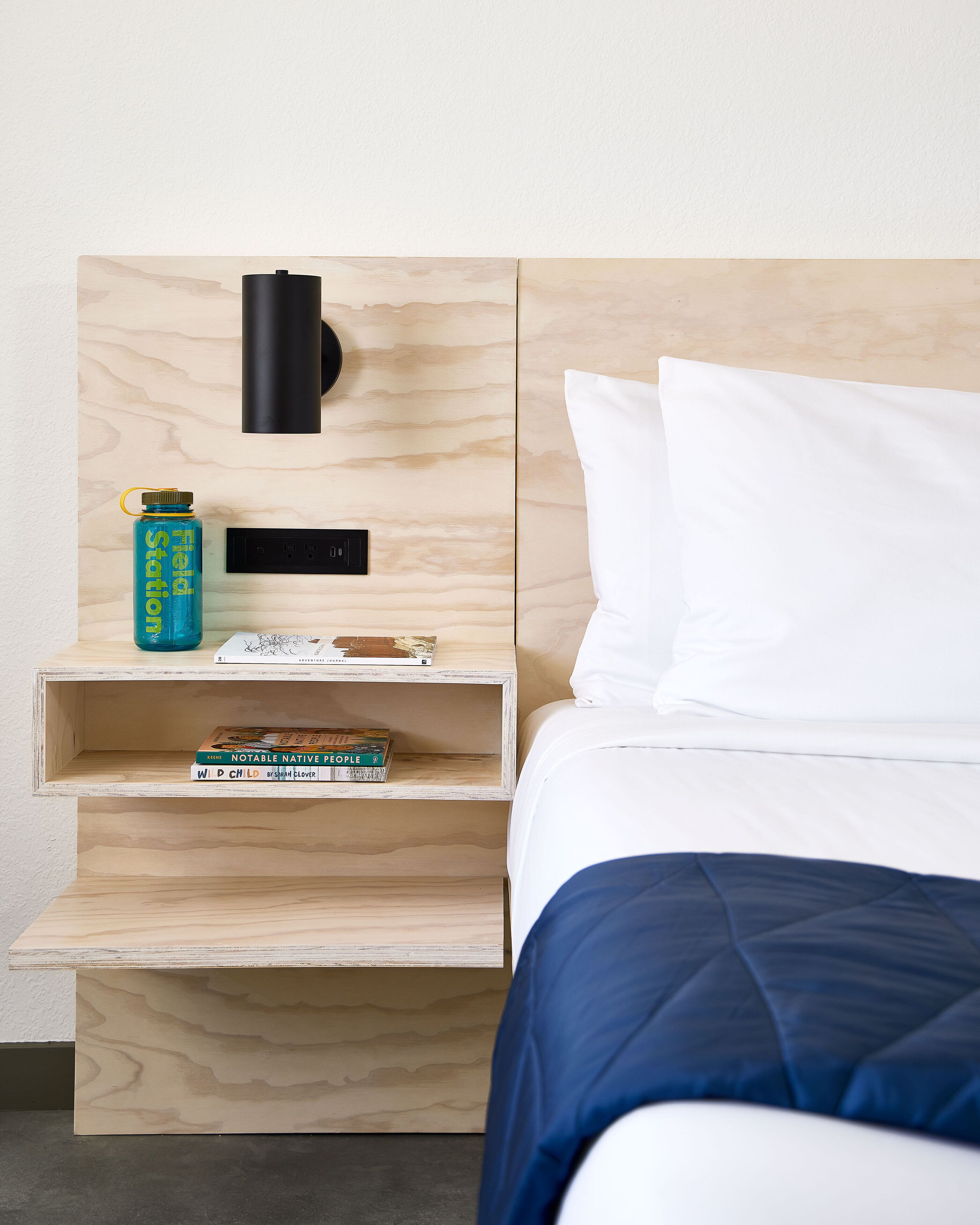 A neatly made bed with white pillows and a blue blanket, a bedside table with a water bottle, a lamp, and two books.