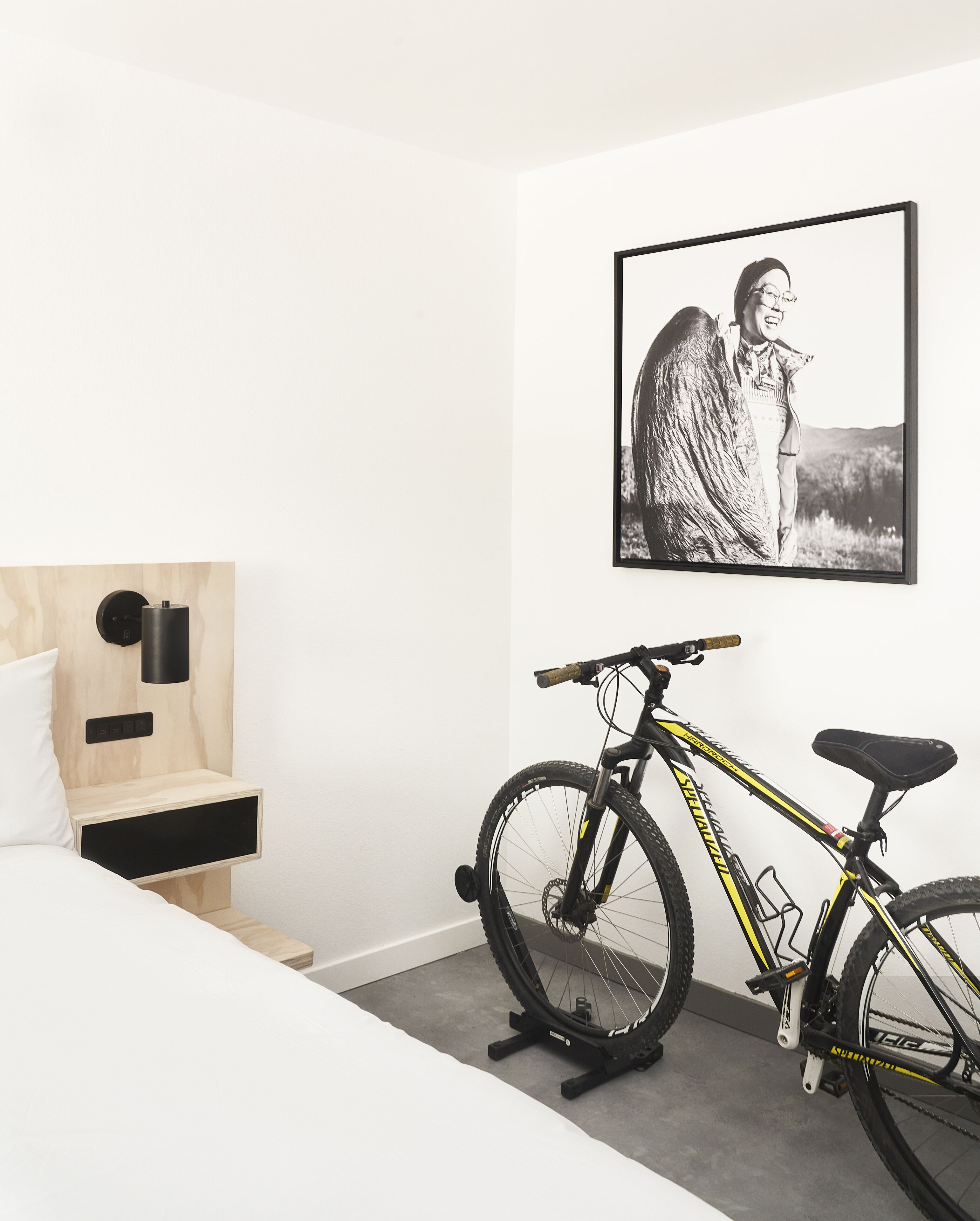 Une chambre à coucher moderne avec un lit, une table de chevet en bois, une lampe fixée au mur, un vélo et un tableau encadré accroché au mur.