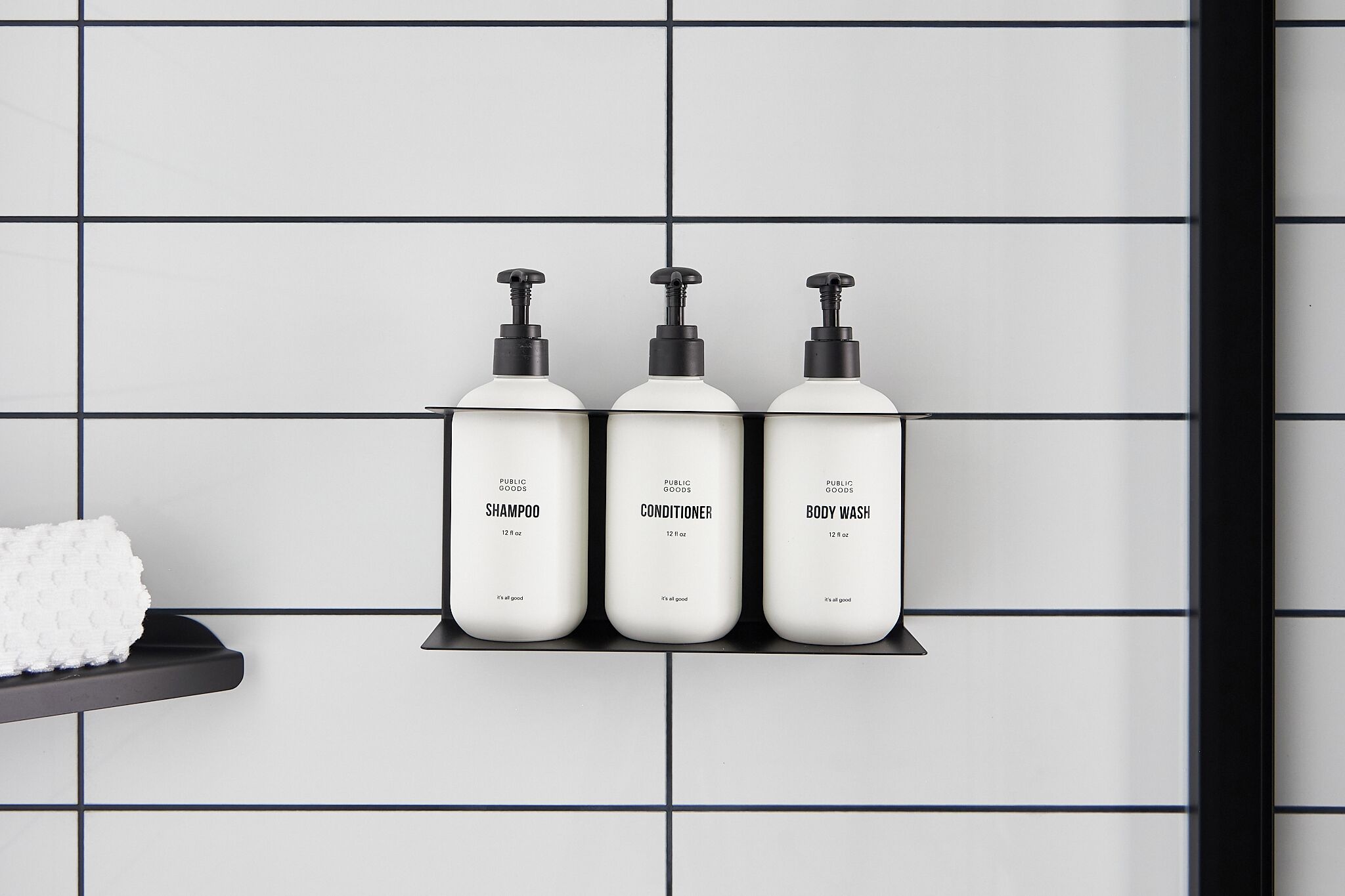 Tres botellas blancas de champú, acondicionador y gel de baño sobre un estante negro frente a una pared con azulejos.