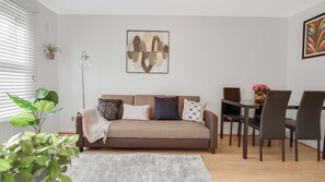 A living room with a brown sofa, a dining table with chairs, a floor lamp, and a potted plant.
