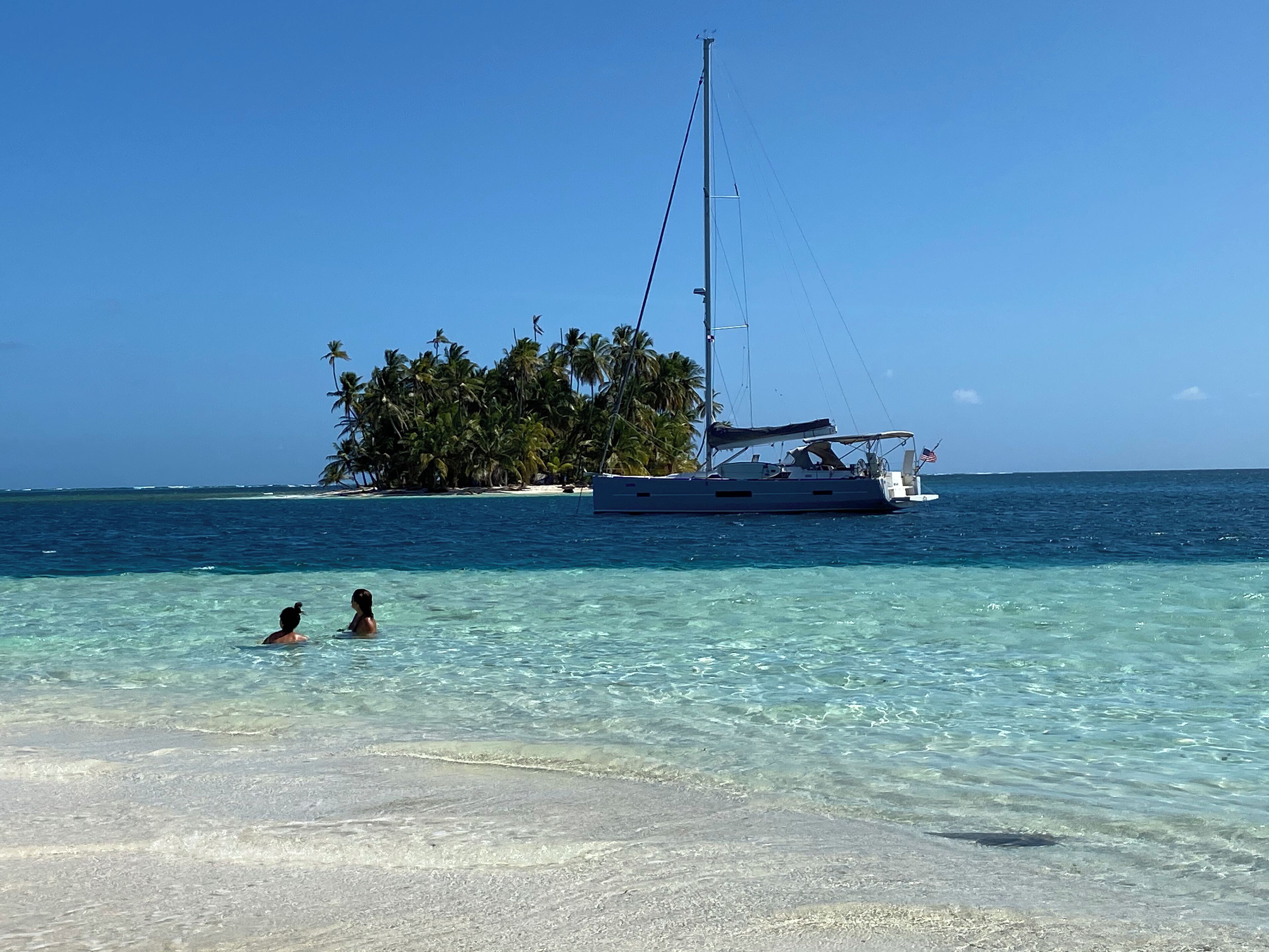 A clear turquoise sea with a sandy beach, a sailboat anchored near a small island with palm trees, and two people swimming in the water.