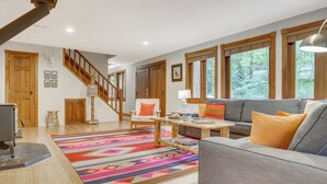 A spacious living room with a gray sofa, colorful rug, wooden staircase, and large windows offering a view of the outdoors. - Mt. Hood Alpine Chalet w/ hot tub, kid & dog friendly (Rhododendron)