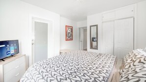 Hotellihuone, jossa on sänky, jalustalla oleva televisio, ovi ja peili seinällä.