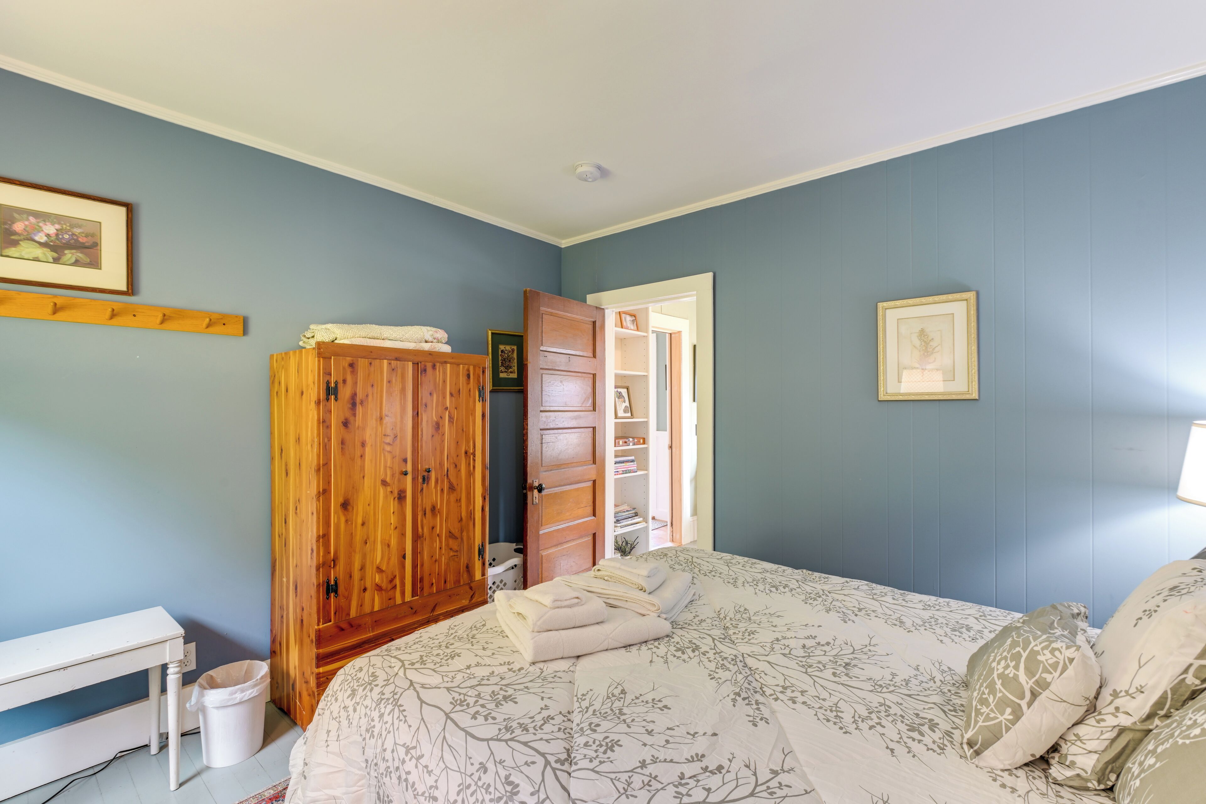A bedroom with a bed, a wooden wardrobe, a white desk, and a small trash bin.