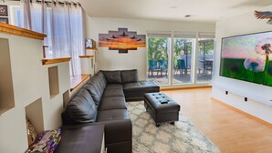 A modern living room with a sectional sofa, a flat-screen TV, and a large window with a view of the outdoors. - Family & Business • Minutes to Spokane Airport, • Fairchild AFB & Northern Quest (Spokane)