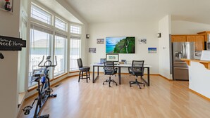 A spacious living area with a large window, a dining table with chairs, an exercise bike, a kitchen area with a refrigerator, and a flat-screen TV mounted on the wall. - Family & Business • Minutes to Spokane Airport, • Fairchild AFB & Northern Quest (Spokane)