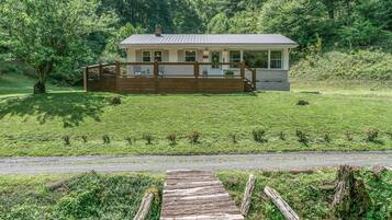A house with a wooden deck, surrounded by a grassy field and trees.