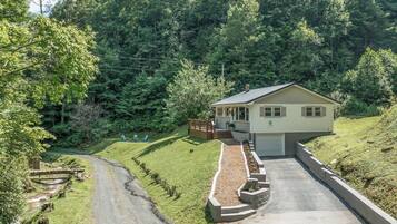 A single-story house with a paved driveway leading to a garage, surrounded by a well-maintained lawn and dense forest.