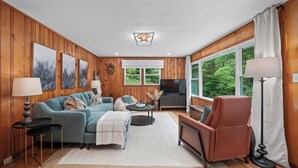 A modern living room with wooden paneling, a large TV, and a view of greenery through the windows. - Charming Mtn Cabin, Hot Tub+Fire Pit, Private, WiFi, Grill, Near Green Mtn Area! (Green Mountain)