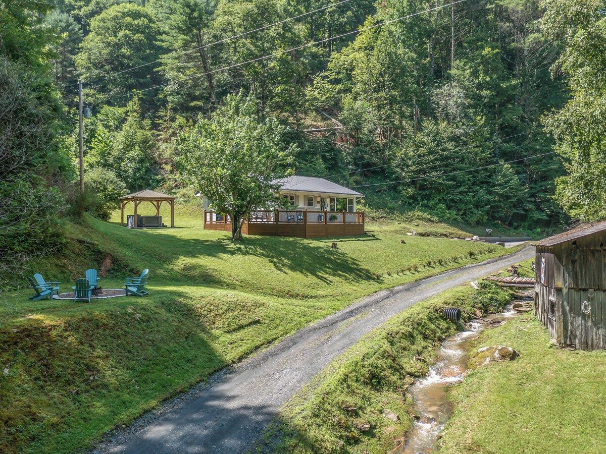 A cabin with a porch, a small outdoor seating area with chairs, and a stream running alongside a road.