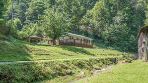 A rustic cabin with a wooden deck and a covered picnic area, surrounded by lush greenery and a small stream. - Charming Mtn Cabin, Hot Tub+Fire Pit, Private, WiFi, Grill, Near Green Mtn Area! (Green Mountain)