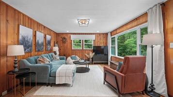 A cozy living room with wooden paneled walls, a large window, a flat-screen TV, and a fireplace.