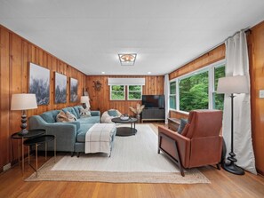 A cozy living room with wooden paneled walls, a large window, a flat-screen TV, and a fireplace.