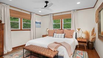 A bedroom with a bed, bedside tables, a bench, a ceiling fan, and windows with curtains.