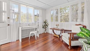 Ein Zimmer mit Holzboden, einem weißen Schreibtisch, einem weißen Stuhl, einem hölzernen Stuhl, einem kleinen Tisch mit einer Vase und einer Topfpflanze.