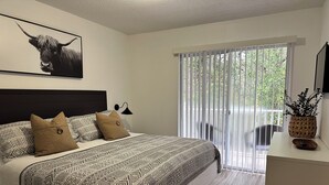 A bedroom with a bed, patterned bedding, a mounted artwork, a bedside lamp, a window with blinds, and a small table with a plant and a phone. - Modern Disney Retreat! 2 Pools + Prime location (Kissimmee)