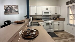 A modern kitchen with white cabinets, a stainless steel sink, and a large island with a tray of mugs. - Modern Disney Retreat! 2 Pools + Prime location (Kissimmee)