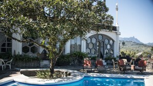 Ein Swimmingpool mit Baum im Vordergrund, umgeben von Sitzgelegenheiten im Freien und einem Gebäude mit markanter Architektur.