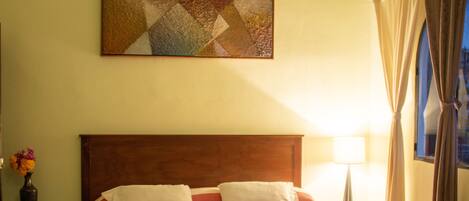 A hotel room with a wooden headboard, a large bed with striped bedding, a decorative painting above the bed, a nightstand with a lamp, and a window with curtains.