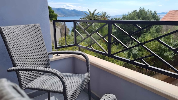 Een balkon met een rieten stoel, een metalen hekwerk en uitzicht op bergen en bomen.