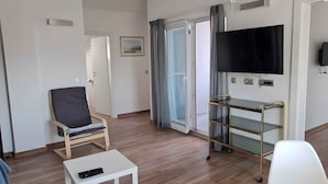 Een moderne woonkamer met een flatscreen televisie, een glazen salontafel, een comfortabele fauteuil en een schuifpui die uitkomt op een balkon.