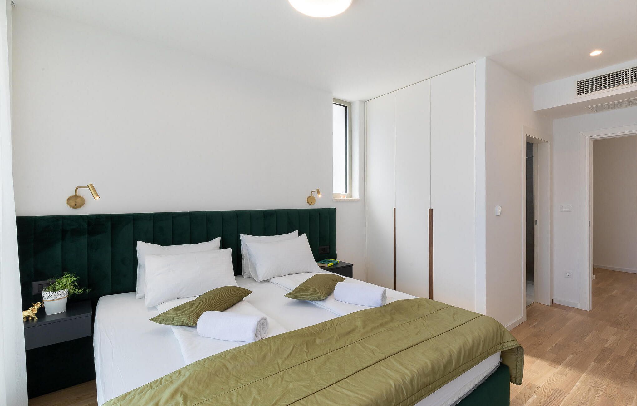 A neatly made bed with white linens and a green throw, a bedside table with a plant, and a window providing natural light.