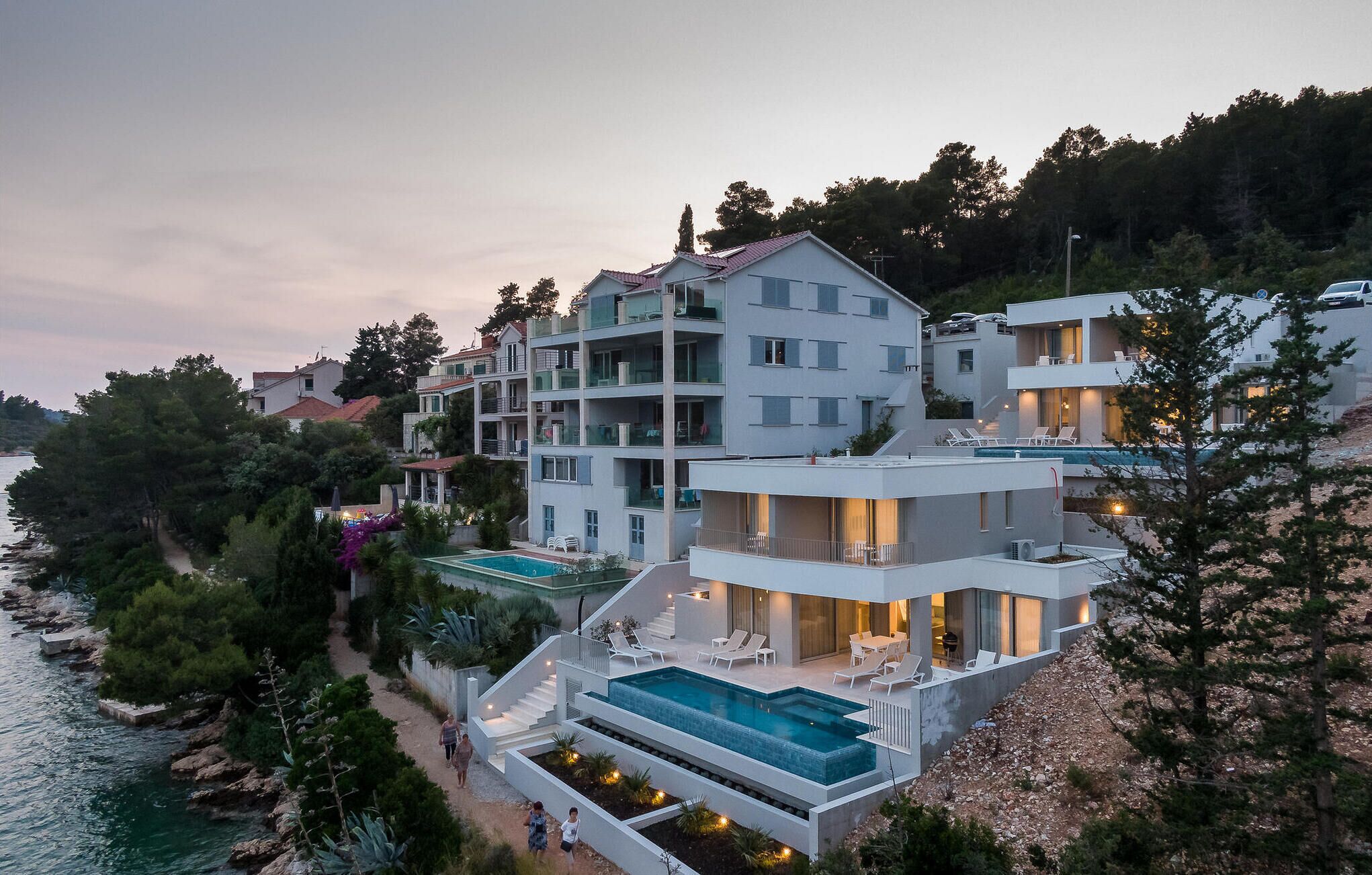 A modern multi-story building with a rooftop pool and outdoor seating area, surrounded by trees and overlooking a body of water.