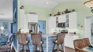 A kitchen with white cabinets, a stainless steel refrigerator, and a microwave. There are wicker chairs around a small island with a sink. - South Gulf Cove 3 Bedroom Canal Pool Home (Port Charlotte)