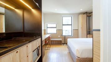 A compact hotel room with a kitchenette, a bed, a small table, and two windows.