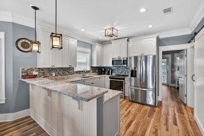 Una cocina moderna con armarios blancos, una isla con encimera de granito, electrodomésticos de acero inoxidable e iluminación colgante.