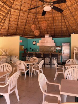 Enjoy dining in our restaurant with a thatched roof ceiling and central bar area, featuring tables and chairs for a comfortable meal.