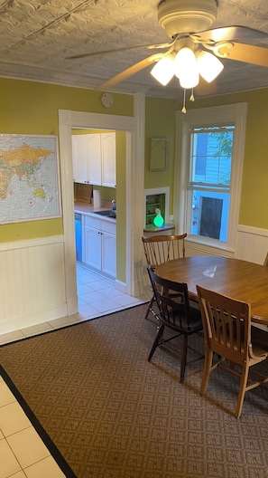 A dining area with a wooden table and chairs, a map on the wall, a kitchen in the background, and a window with blinds.
