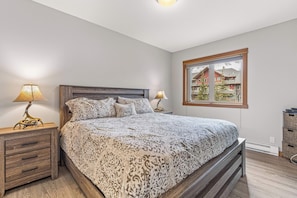 A bedroom with a large bed, two bedside lamps, a wooden nightstand, and a window with a view of houses.