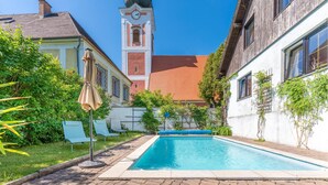 A swimming pool with a brick pathway, surrounded by greenery and buildings with a clock tower.