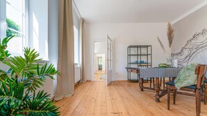 Une pièce avec un plancher en bois, une table à manger avec des chaises, une bibliothèque et une grande fenêtre avec des rideaux.