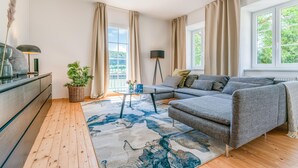 Un salon moderne avec un canapé d’angle gris, un plancher en bois, un tapis à motifs, un lampadaire et une vue sur la verdure grâce à de grandes fenêtres.