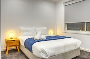 A neatly made bed with white linens and a blue throw blanket, a bedside table with a lamp, and a window with a blind. - Bella - Modern, two bedroom townhouse close to Ballarat CBD (Bakery Hill)