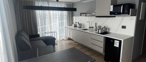Een moderne keuken met een magnetron, spoelbak en kastjes. Er is een eettafel met stoelen en een bank in de woonkamer.