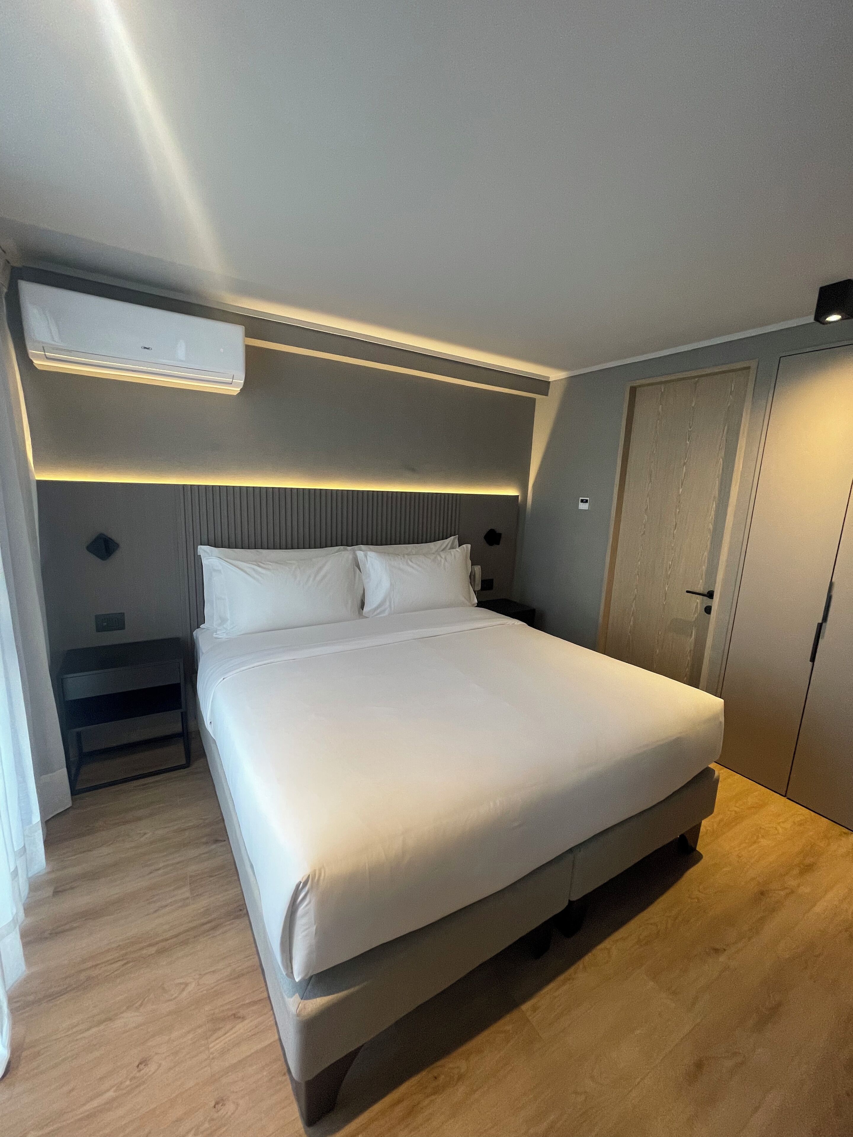 A modern bedroom with a large bed, a bedside table, a wardrobe, and an air conditioning unit.