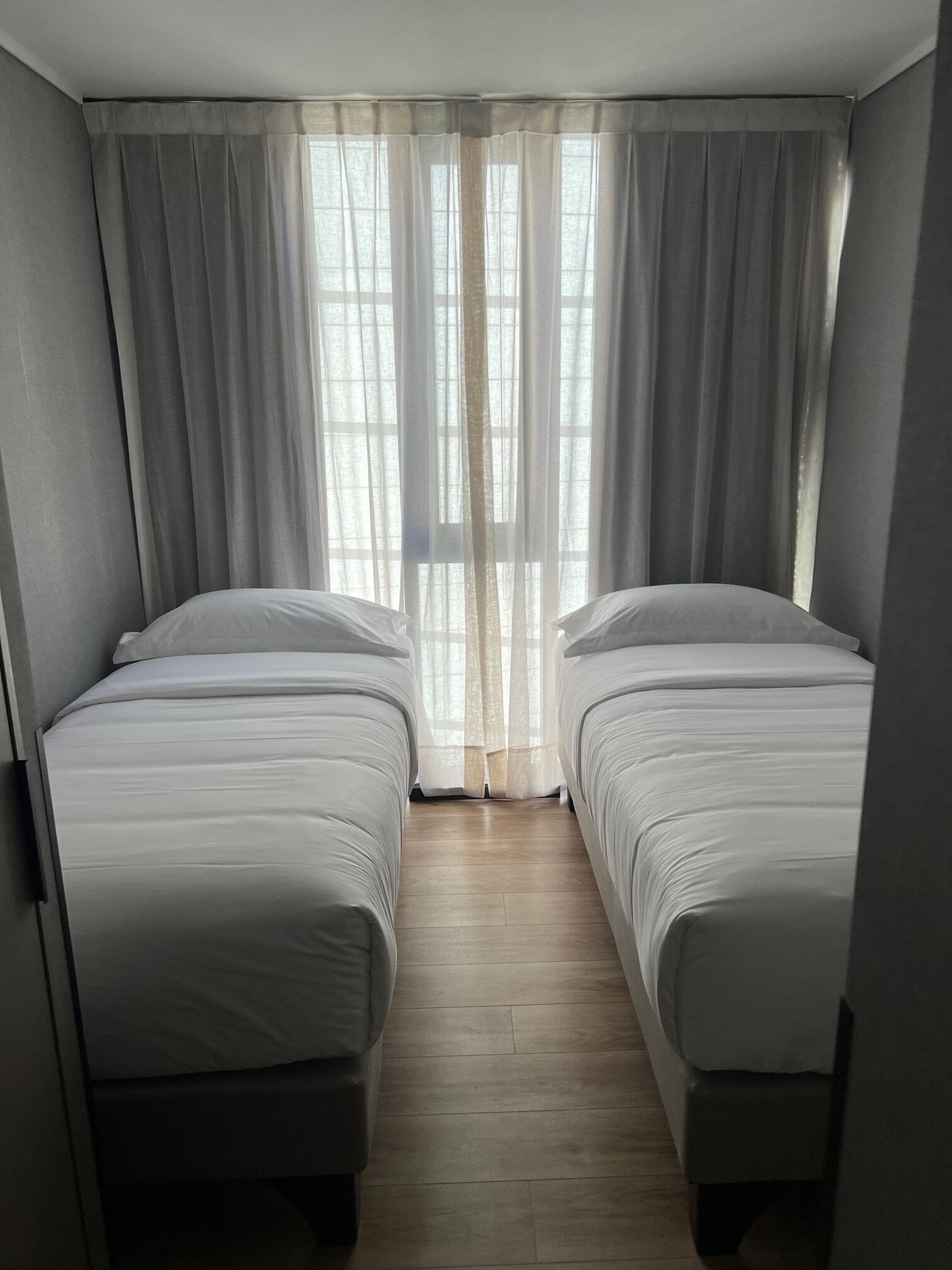 A room with two single beds, a wooden floor, and a window with sheer curtains.