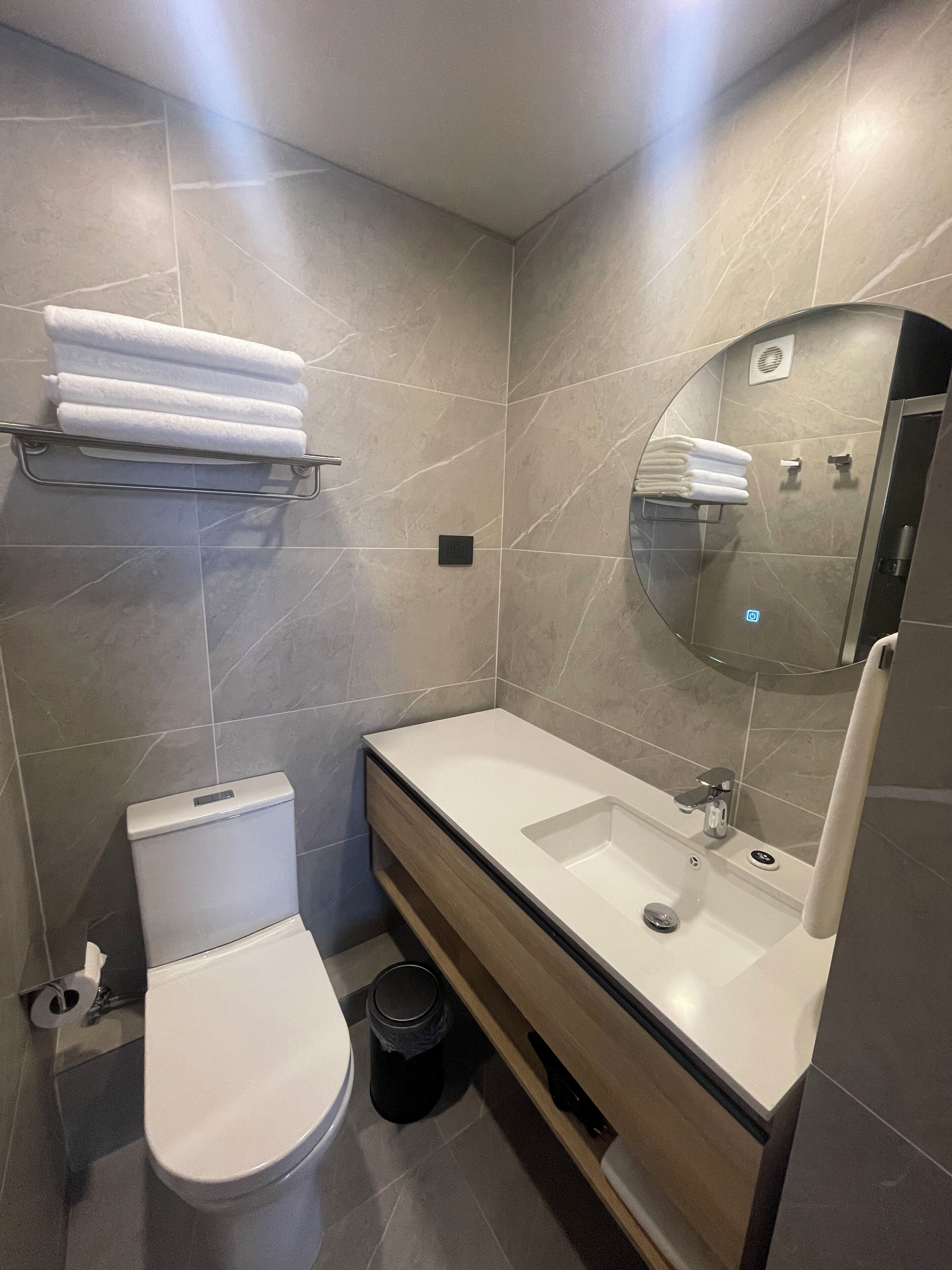 A modern bathroom with a toilet, sink, and mirror. There are towels on a shelf and a trash bin.