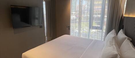 A modern hotel room with a large bed, a flat-screen TV mounted on the wall, and a window with sheer curtains.