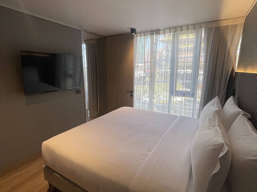 A modern hotel room with a large bed, a flat-screen TV mounted on the wall, and a window with sheer curtains.